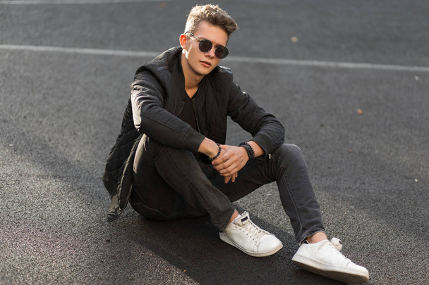 Νεαρό όμορφος hipster άντρας με γυαλιά ηλίου με μοντέρνα ρούχα μαύρη με το σακάκι και κομψό λευκό πάνινα παπούτσια κάθονται στην οδό  - Φωτογραφία, εικόνα