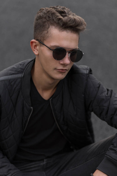 Fashionable handsome young man with stylish vintage sunglasses and black trendy jacket in the street - Фото, зображення