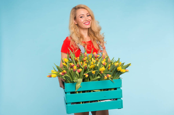 青い背景にチューリップのボックスで陽気な若いブロンドの女性花屋 - 写真・画像