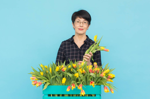Heureuse femme d'âge moyen avec des lunettes tenant boîte de tulipes en fond bleu
 - Photo, image