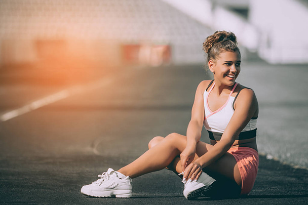 Attractive sporty woman sitting confident on stadium tracks on a sunny summer day. Sports model exercising in summer at morning - Zdjęcie, obraz