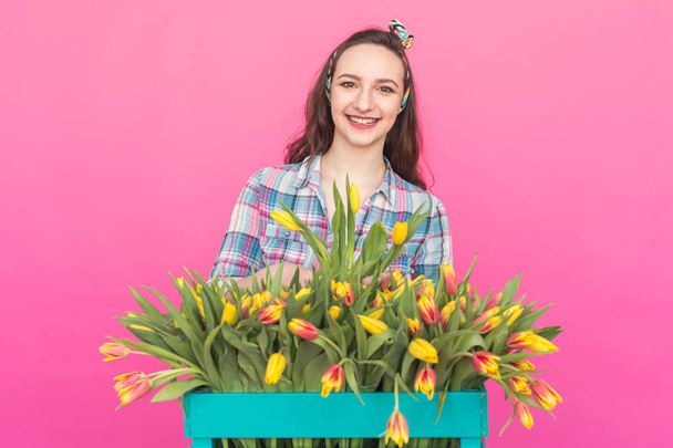 Happy beautiful brunette caucasian young woman with big box of tulips on pink background. - Photo, Image