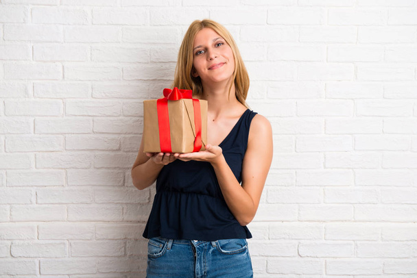 Blondes Mädchen hält ein Geschenk in den Händen auf weißem Backstein-Hintergrund - Foto, Bild