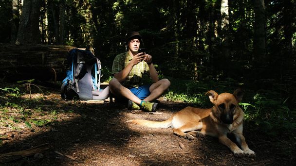 Young man tourist with a dog and a backpack is sitting in the forest and the guy is using a smartphone. - Photo, Image