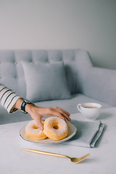 リビング ルーム、家庭での朝の朝食のコンセプトでテーブルのホワイト プレートから砂糖のトッピングで美味しいドーナツを取って手します。  - 写真・画像