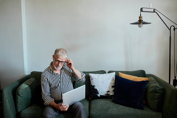 взрослый бородатый мужчина работает из дома с ноутбуком. сидя за столом у окна - Фото, изображение