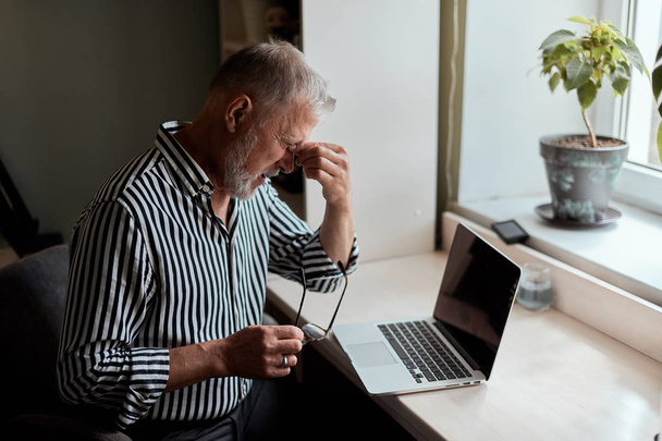 mature man looking exhausted while sitting at his laptop and holding his glasses in hand - Foto, imagen