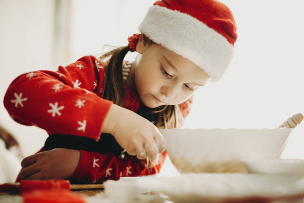 Adorable little girl in Santa hat looking concentrated while preparing Christmas cookies at table - Foto, Bild