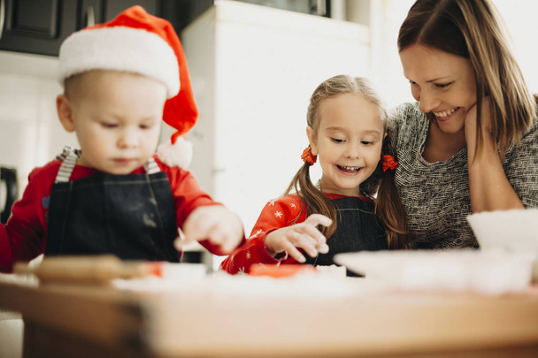 Suloinen pieni poika ja tyttö, jolla on aikuinen hymyilevä äiti pöydässä keittiössä tehden Xmas-evästeitä kaikki yhdessä
 - Valokuva, kuva