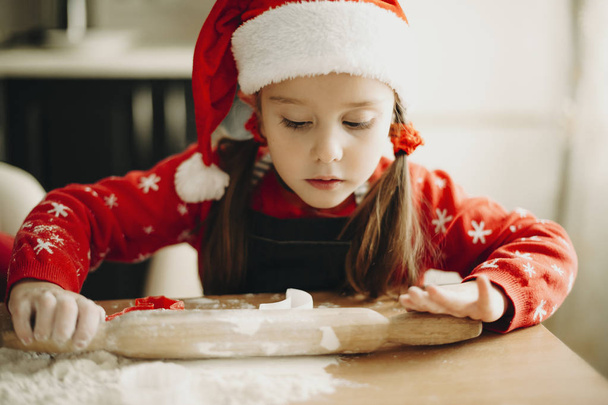 entzückendes konzentriertes Mädchen mit Weihnachtsmütze und mit Nudelholz am Tisch sitzend, um Urlaubsplätzchen zu backen - Foto, Bild