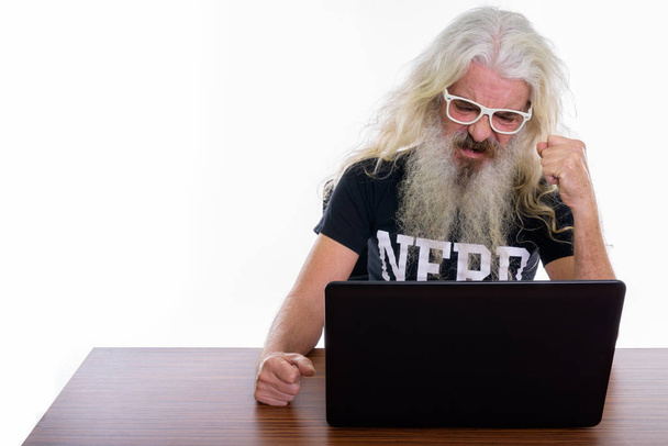 Studio shot of senior barbu nerd homme regardant en colère tout en utilisant un ordinateur portable sur une table en bois - Photo, image
