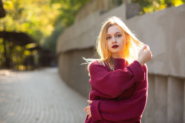Елегантна блондинка з червоними губами носить червоний оверсайз светр, позуючи в променях сонця. Простір для тексту
 - Фото, зображення