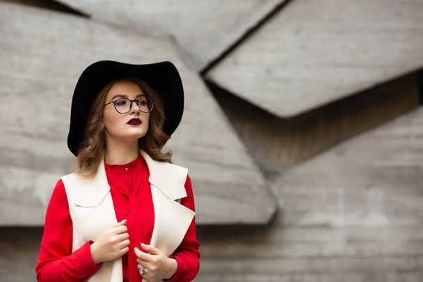 Jovem elegante vestida com lábios vermelhos usando blusa, chapéu e óculos posando na parede de concreto. Espaço para texto
 - Foto, Imagem