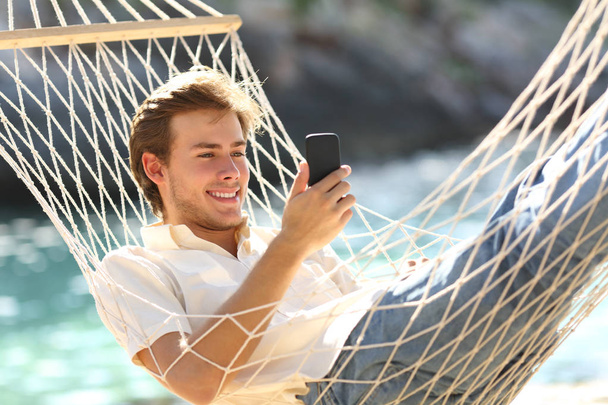 Happy man resting on a hammock using a smart phone on summer bacation on the beach - Photo, Image