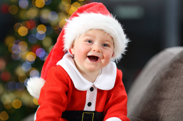 Cheerful baby dressed as santa claus on a couch at home in christmas with a tree in the background - Photo, Image