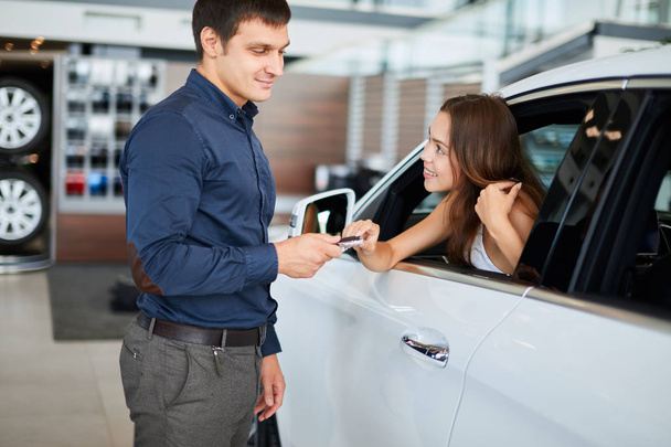  ar salesman giving car keys to young woman sitting in car in auto dealership - Foto, imagen