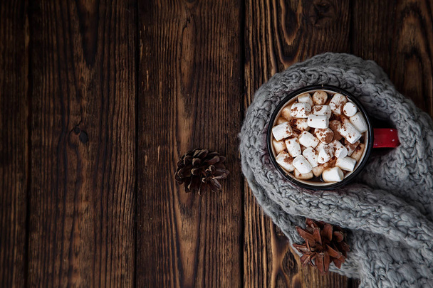 μία κόκκινη κούπα με ζεστό ρόφημα με marshmallow και πλεκτό κασκόλ σε ξύλινο χριστουγεννιάτικο φόντο - Φωτογραφία, εικόνα