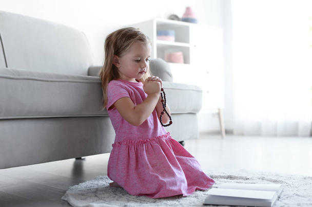 Cute little girl with beads praying over Bible in living room, black and white effect. Space for text - Φωτογραφία, εικόνα