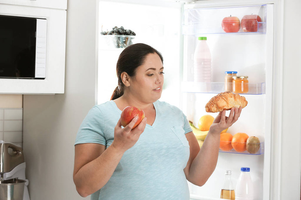 Frau wählt zwischen Apfel und Croissant in der Nähe des Kühlschranks in der Küche. Gesunde Ernährung - Foto, Bild