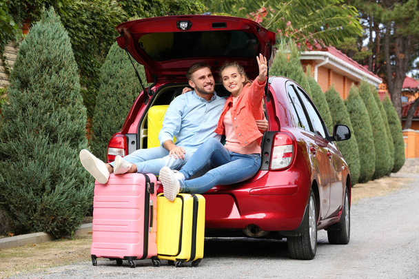 Happy young couple sitting in car trunk and suitcases outdoors - 写真・画像