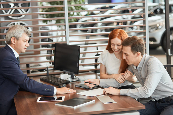 Beautiful young couple signs documents at car dealership office. - 写真・画像