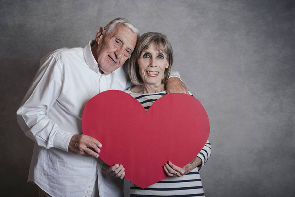 Porträt eines glücklichen Seniorehepaares mit rotem Herz auf grauem Hintergrund - Foto, Bild