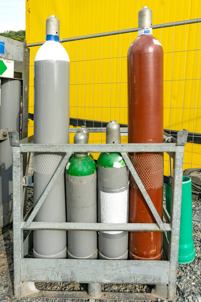 Valikoima erilaisia kaasupulloja metalli seistä ulkona soralla takana kirkkaan keltainen rakenne
 - Valokuva, kuva