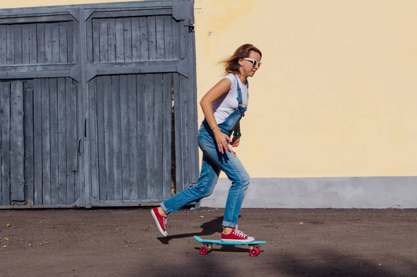 Portrait of a smiling woman dressed in overalls and sunglasses riding on her plastic skateboard next to the old wooden wall. - Valokuva, kuva