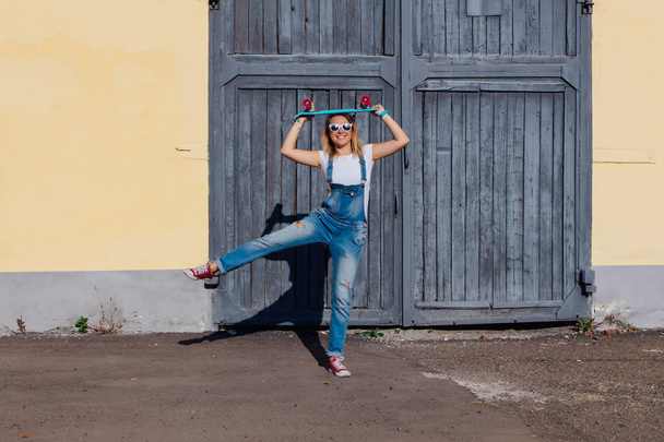 Portrait of a smiling woman dressed in overalls and sunglasses standing with her skateboard next to the old wooden wall. - Фото, изображение