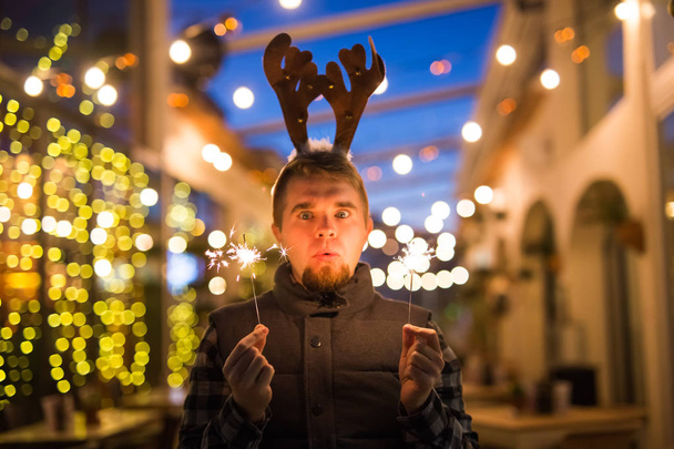 Έννοια διακοπές Χριστουγέννων, άνθρωποι και χειμώνα - έκπληκτος ο άνθρωπος στα κέρατα του ελαφιού στέκεται στο δρόμο το βράδυ με Βεγγάλη φώτα στα χέρια του - Φωτογραφία, εικόνα