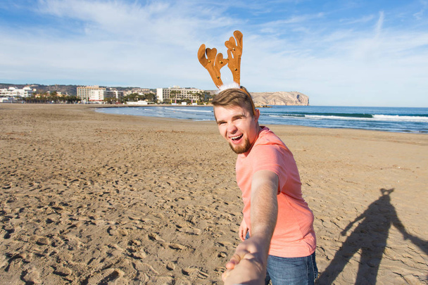 Follow me, holidays and Christmas concept - Happy young man with deer horns holding Santas hand on the beach. - Photo, Image