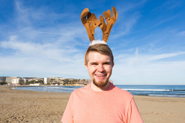 Διασκέδαση, τις διακοπές και ακολούθησέ με έννοια - ευτυχισμένος άνθρωπος στην κέρατα ελαφιών Χριστούγεννα στην παραλία. - Φωτογραφία, εικόνα