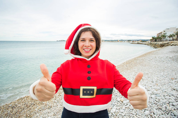 Menschen, Feiertage und Weihnachten - fröhliche Frau mit Weihnachtsmütze zeigt Daumen hoch - Foto, Bild