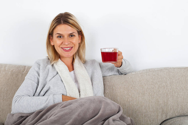 Beautiful woman relaxing on the sofa and drinking hot tea, wrapped in a grey blanket - Foto, Imagem