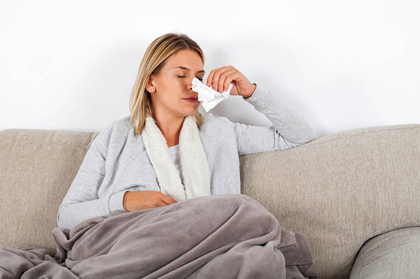 Picture of a sick young lady sitting on the couch and blowing her runny nose - Foto, Bild