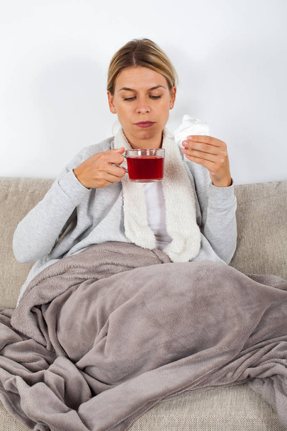 Sick young woman resting on the sofa while drinking hot tea and holding napkins - Foto, Bild