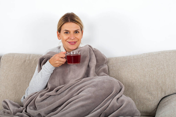 Sick young woman resting on the sofa while drinking hot tea and holding napkins - Photo, Image