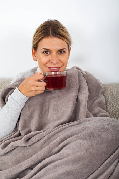 Mujer joven enferma descansando en el sofá mientras bebe té caliente y sostiene servilletas
 - Foto, Imagen