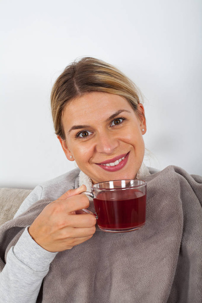 Sick young woman resting on the sofa while drinking hot tea and holding napkins - Foto, Imagen