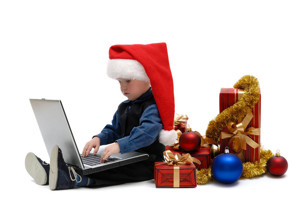 Little boy in Santa Claus cap with a laptop and Christmas gifts on white background, isolated - Fotoğraf, Görsel