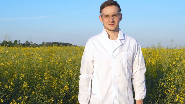 Portrait of a young handsome biologist or agronomist in white medical clothes, safety glasses, smiling, successful, field of canola, bio. - 写真・画像