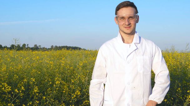 Portrait of a young handsome biologist or agronomist in white medical clothes, safety glasses, smiling, successful, field of canola, bio. - Foto, immagini