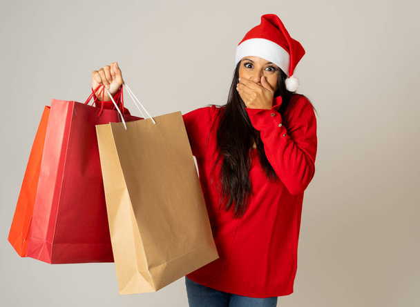 Young woman ready for christmas with paper shopping bags in red and Santa claus hat isolated grey background in being ready for christmas shopping gifts celebration consumerism and sales concept. - Foto, afbeelding