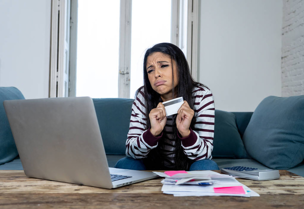 Attractive young latin woman desperate and furious with credit card and laptop siting on sofa home in credit card problems debts online payment online shopping online banking bad financial situation. - Photo, Image