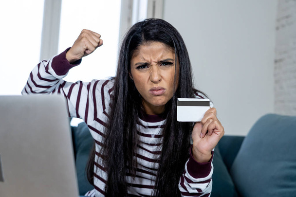 Attractive young latin woman desperate and furious with credit card and laptop siting on sofa home in credit card problems debts online payment online shopping online banking bad financial situation. - Foto, afbeelding