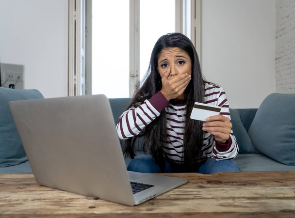 Attractive young latin woman desperate and furious with credit card and laptop siting on sofa home in credit card problems debts online payment online shopping online banking bad financial situation. - Photo, image