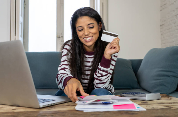Cheerful attractive latin woman using credit card calculator and laptop paying bills at home looking relax in home finances paying bills online banking buying online and internet shopping. - Photo, Image
