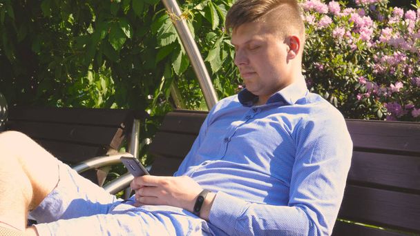 Young handsome student guy is sitting in the park on a bench, mobile phone. Concept: communication - Valokuva, kuva