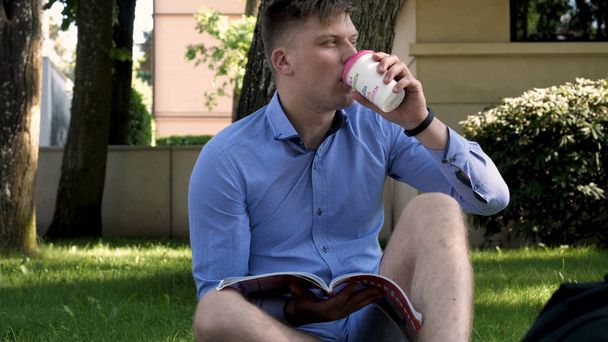 Young handsome student guy sitting in the park, writes messages in the phone and drinks coffee. Concept: study, rest, communication, chat. - Фото, зображення