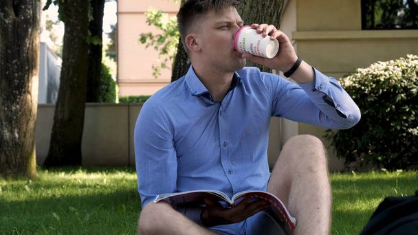 Young handsome student guy sitting in the park, writes messages in the phone and drinks coffee. Concept: study, rest, communication, chat. - Fotografie, Obrázek